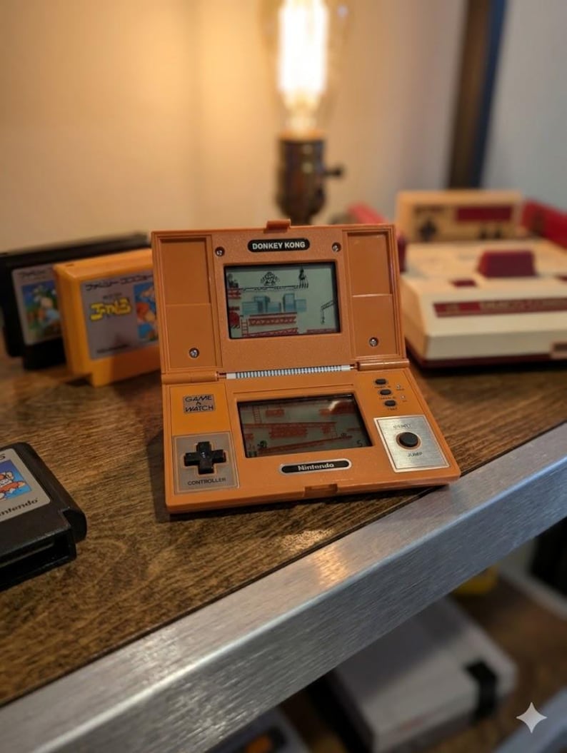 K&ouml;nnte beinhalten: Orangefarbene Nintendo Game & Watch Handheld-Konsole mit dem Spiel Donkey Kong. Die Konsole hat einen kreuzf&ouml;rmigen Controller und eine Starttaste. Mehrere Spielkassetten und eine klassische Nintendo-Konsole sind im Hintergrund zu sehen.