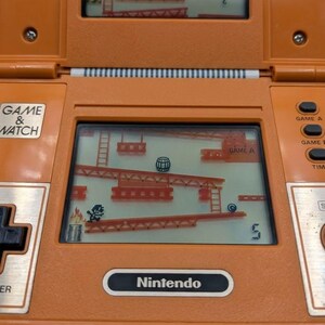 K&ouml;nnte beinhalten: Orangefarbenes Nintendo Game & Watch Handheld-Elektronikspiel. Der Bildschirm zeigt ein klassisches Arcade-Spiel mit einer Figur, die Leitern erklimmt, um einem rollenden Fass auszuweichen. Das Spiel hat Tasten f&uuml;r Spiel A, Spiel B und Zeit.
