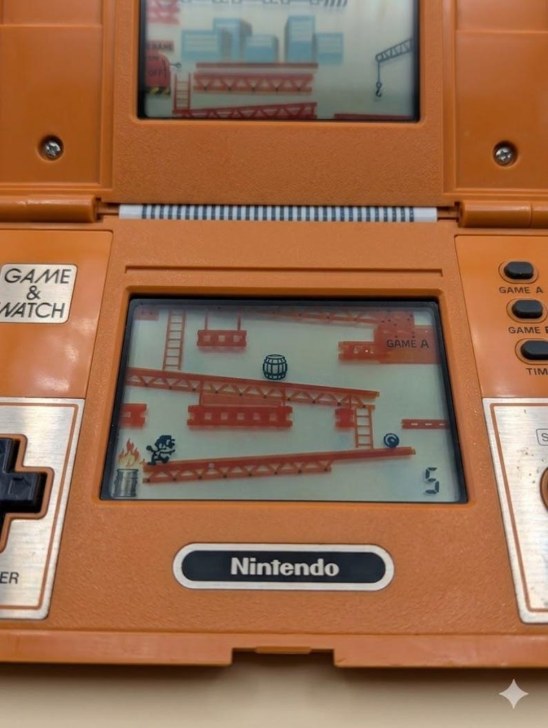 K&ouml;nnte beinhalten: Orangefarbenes Nintendo Game & Watch Handheld-Elektronikspiel. Der Bildschirm zeigt ein klassisches Arcade-Spiel mit einer Figur, die Leitern erklimmt, um einem rollenden Fass auszuweichen. Das Spiel hat Tasten f&uuml;r Spiel A, Spiel B und Zeit.