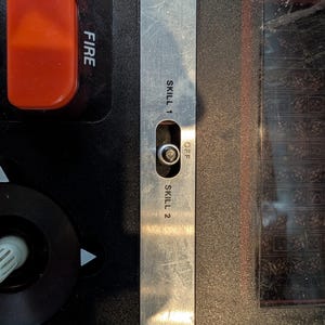 May include: Close-up of a control panel with a silver metal switch labeled "SKILL 1", "OFF", and "SKILL 2". A red button labeled "FIRE" is visible. The panel is black and orange.