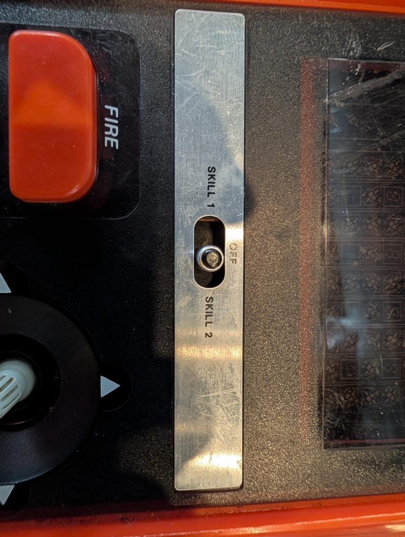 May include: Close-up of a control panel with a silver metal switch labeled "SKILL 1", "OFF", and "SKILL 2". A red button labeled "FIRE" is visible. The panel is black and orange.