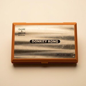 K&ouml;nnte beinhalten: Ein Vintage Nintendo Game & Watch Donkey Kong Handheld-Spiel. Das Ger&auml;t hat einen orangefarbenen Rahmen und einen silbernen Bildschirm mit dem Spieltitel. In der oberen linken Ecke steht "Game & Watch" und "Nintendo". Unten steht "Multi Screen".