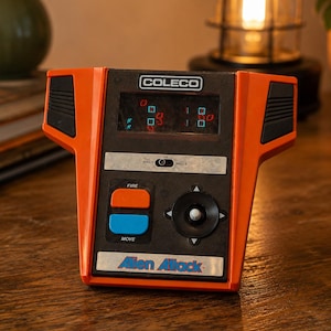 May include: Vintage Coleco Alien Attack handheld game. The game is orange and black with a small screen displaying pixelated graphics. It has buttons labeled "FIRE" and "MOVE", and the game's title is printed on the front.