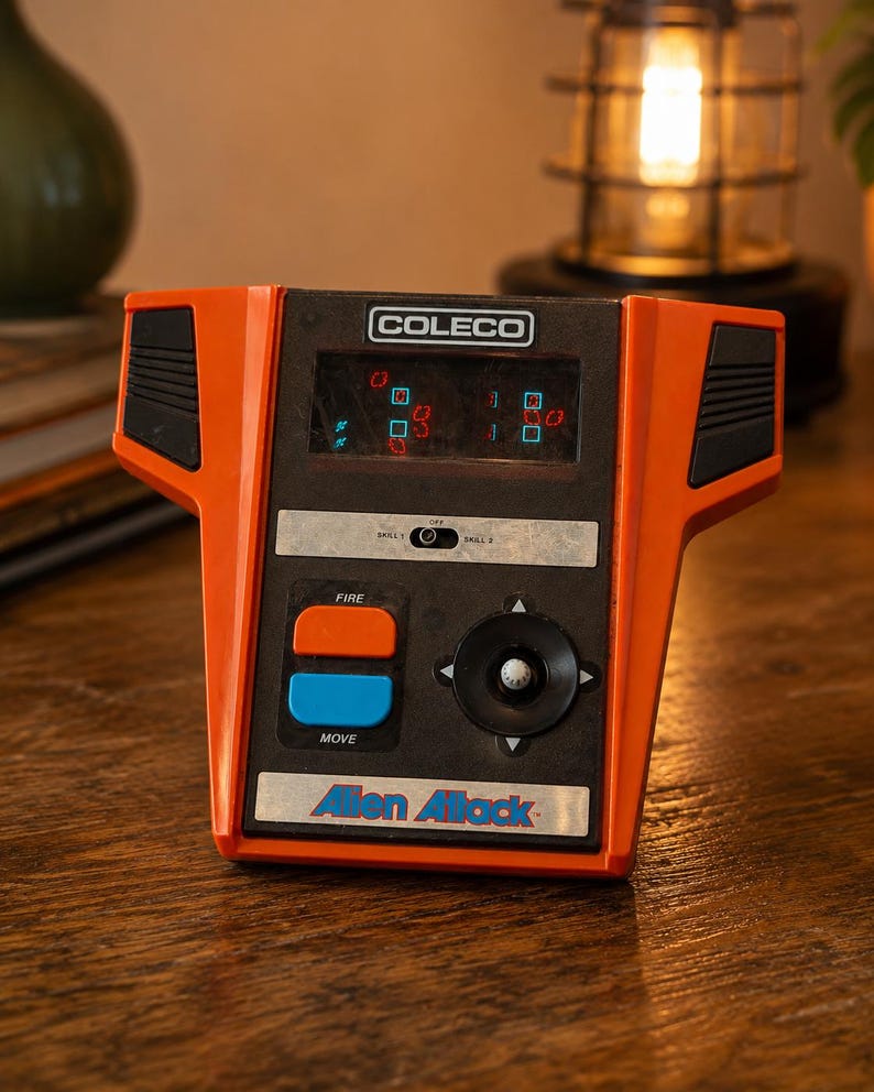 May include: Vintage Coleco Alien Attack handheld game. The game is orange and black with a small screen displaying pixelated graphics. It has buttons labeled "FIRE" and "MOVE", and the game's title is printed on the front.