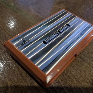 K&ouml;nnte beinhalten: Ein Vintage Donkey Kong Multi Screen Handheld-Spiel. Das Spiel hat eine silberne Oberfl&auml;che mit dem Spieltitel in Schwarz und einen braunen Kunststoffrahmen. Die W&ouml;rter "Game & Watch" und "Nintendo" sind ebenfalls sichtbar.