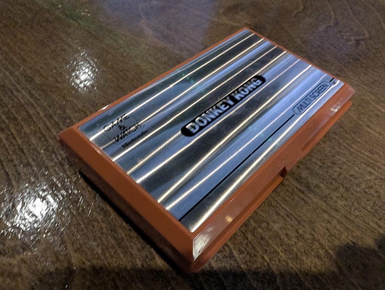K&ouml;nnte beinhalten: Ein Vintage Donkey Kong Multi Screen Handheld-Spiel. Das Spiel hat eine silberne Oberfl&auml;che mit dem Spieltitel in Schwarz und einen braunen Kunststoffrahmen. Die W&ouml;rter "Game & Watch" und "Nintendo" sind ebenfalls sichtbar.