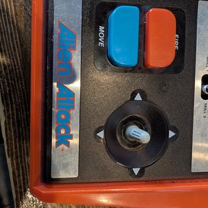 May include: Close-up of a vintage "Alien Attack" game console control panel. The panel is primarily black and orange, featuring a joystick, blue and red buttons labeled "MOVE" and "FIRE", and the game's logo.