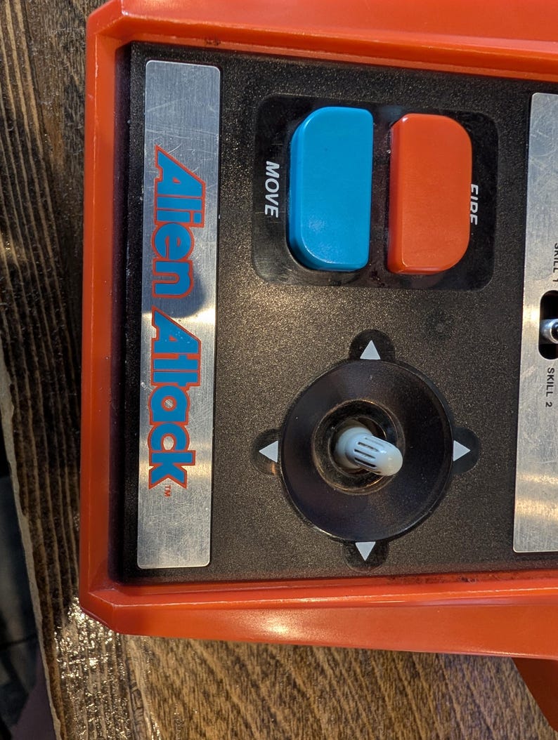 May include: Close-up of a vintage "Alien Attack" game console control panel. The panel is primarily black and orange, featuring a joystick, blue and red buttons labeled "MOVE" and "FIRE", and the game's logo.