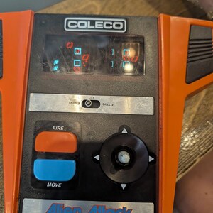 May include: An orange and black Coleco Alien Attack handheld video game. The game features a screen displaying pixelated graphics, a directional pad, and buttons labeled "FIRE" and "MOVE". The game has a skill level selector.