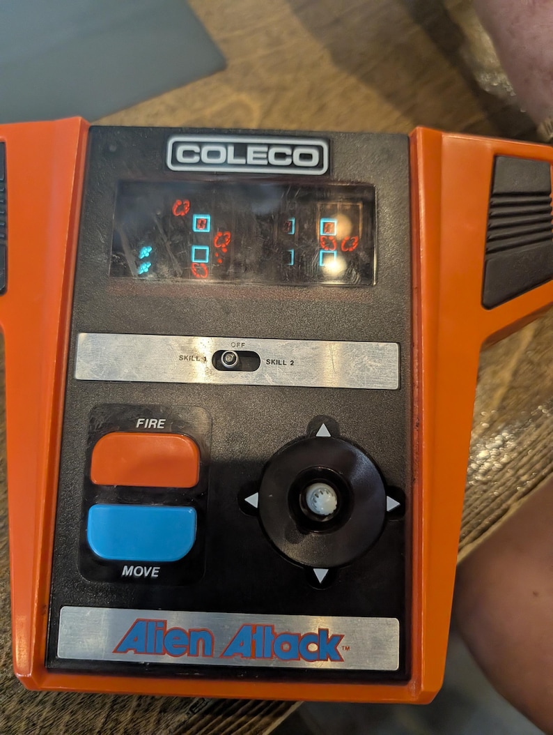 May include: An orange and black Coleco Alien Attack handheld video game. The game features a screen displaying pixelated graphics, a directional pad, and buttons labeled "FIRE" and "MOVE". The game has a skill level selector.