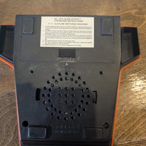 May include: A black and orange Coleco Alien Attack video game console. The console has a speaker grill, a battery compartment, and a label with operating instructions. The text on the label includes "Alien Attack" and "4 C Alkaline Batteries Required."