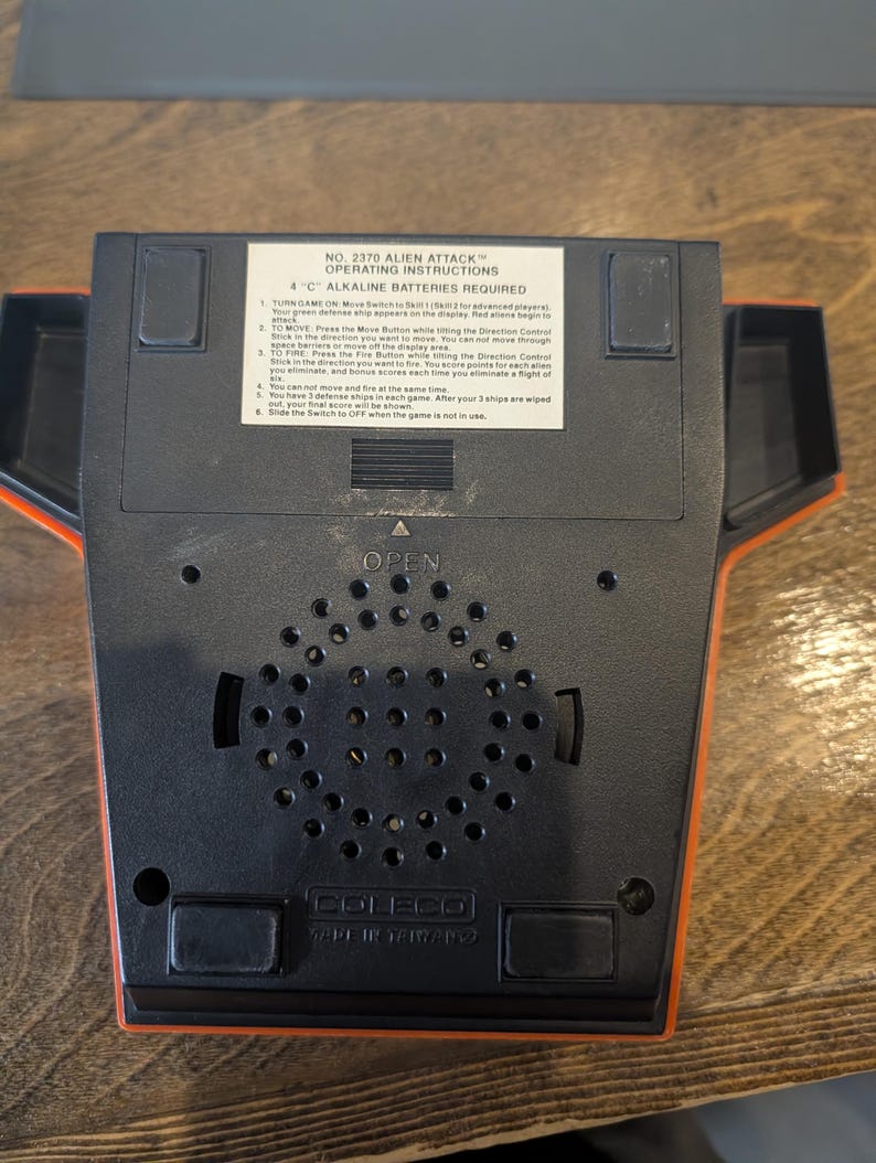 May include: A black and orange Coleco Alien Attack video game console. The console has a speaker grill, a battery compartment, and a label with operating instructions. The text on the label includes "Alien Attack" and "4 C Alkaline Batteries Required."