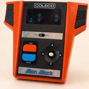 May include: Vintage Coleco Alien Attack handheld game. The game features a black and orange design with a directional pad, fire and move buttons, and a small screen. The game's logo is visible at the bottom.