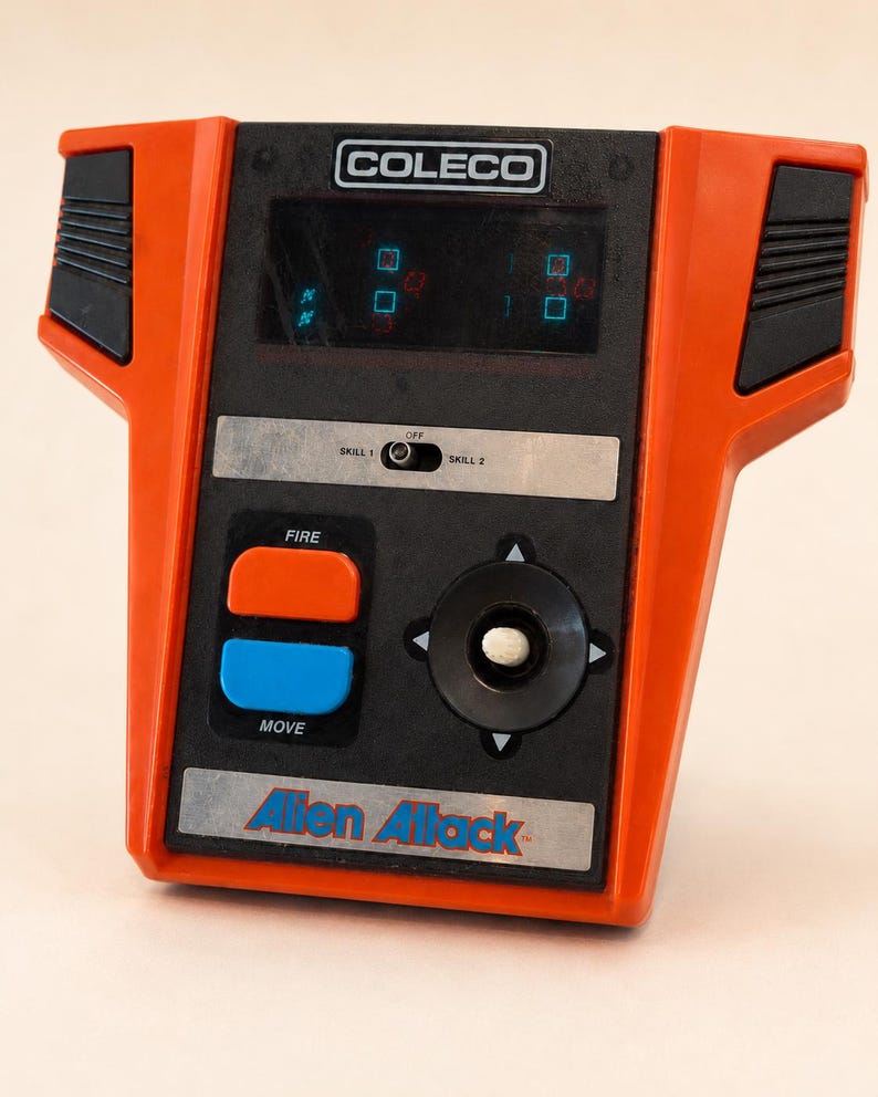 May include: Vintage Coleco Alien Attack handheld game. The game features a black and orange design with a directional pad, fire and move buttons, and a small screen. The game's logo is visible at the bottom.