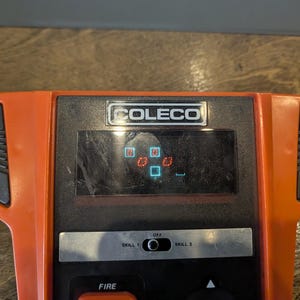 May include: An orange and black Coleco handheld video game console. The screen displays a game in progress with red and blue block shapes. The console has a black control panel with a "FIRE" button and a "SKILL 1" and "SKILL 2" switch.