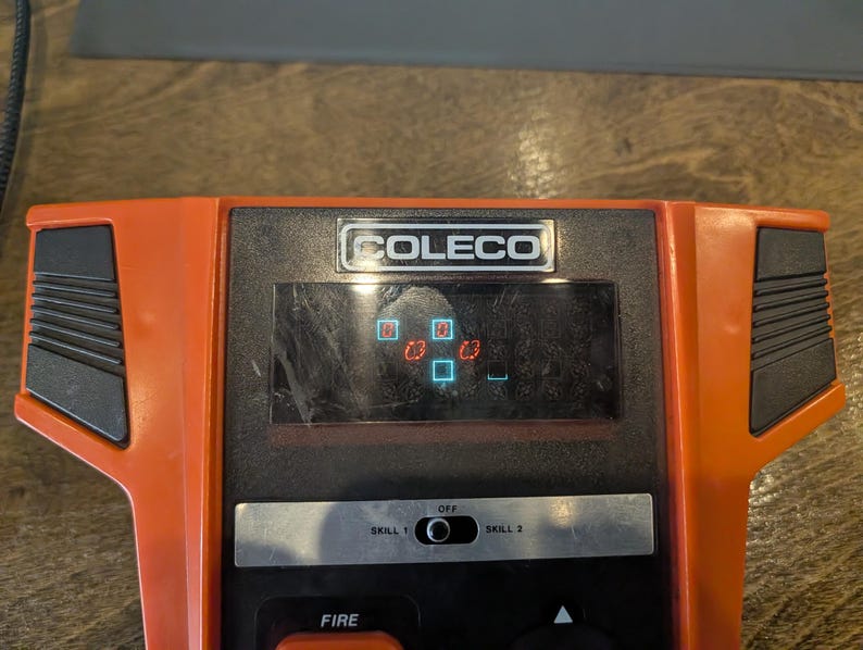 May include: An orange and black Coleco handheld video game console. The screen displays a game in progress with red and blue block shapes. The console has a black control panel with a "FIRE" button and a "SKILL 1" and "SKILL 2" switch.