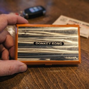 K&ouml;nnte beinhalten: Ein orangefarbenes und silbernes Nintendo Game & Watch Donkey Kong Multi Screen Handheld-Spiel. Das Spiel ist rechteckig mit dem Donkey Kong-Logo in der Mitte. Das Spiel liegt auf einer Holzoberfl&auml;che.