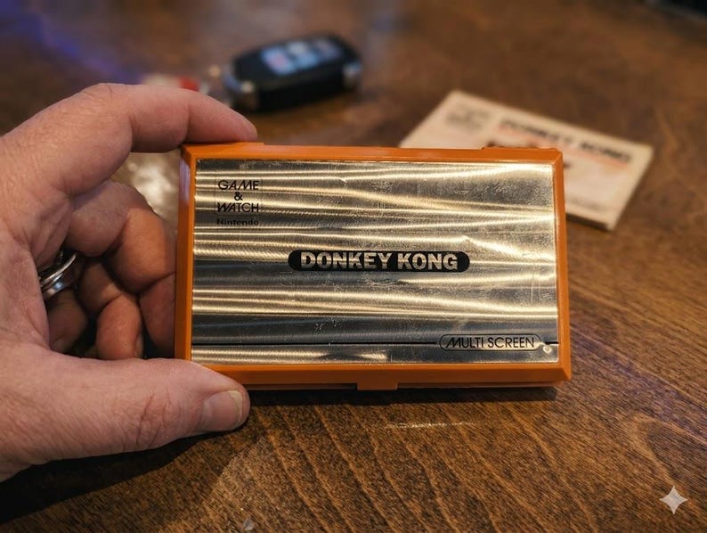 K&ouml;nnte beinhalten: Ein orangefarbenes und silbernes Nintendo Game & Watch Donkey Kong Multi Screen Handheld-Spiel. Das Spiel ist rechteckig mit dem Donkey Kong-Logo in der Mitte. Das Spiel liegt auf einer Holzoberfl&auml;che.
