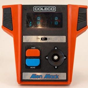 May include: Vintage Coleco tabletop arcade game, "Alien Attack." The game features a black and orange design with a screen displaying pixelated graphics. It has buttons labeled "FIRE" and "MOVE," a joystick, and the Coleco logo.