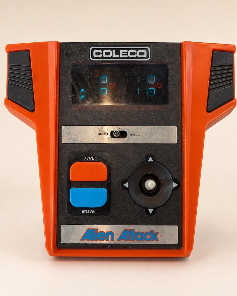 May include: Vintage Coleco tabletop arcade game, "Alien Attack." The game features a black and orange design with a screen displaying pixelated graphics. It has buttons labeled "FIRE" and "MOVE," a joystick, and the Coleco logo.