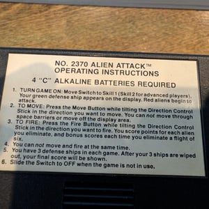 May include: A close-up of a black game console with a white label. The label reads "NO. 2370 ALIEN ATTACK" and includes operating instructions. The text details how to play the game, including moving, firing, and game rules. Requires 4 "C" alkaline batteries.
