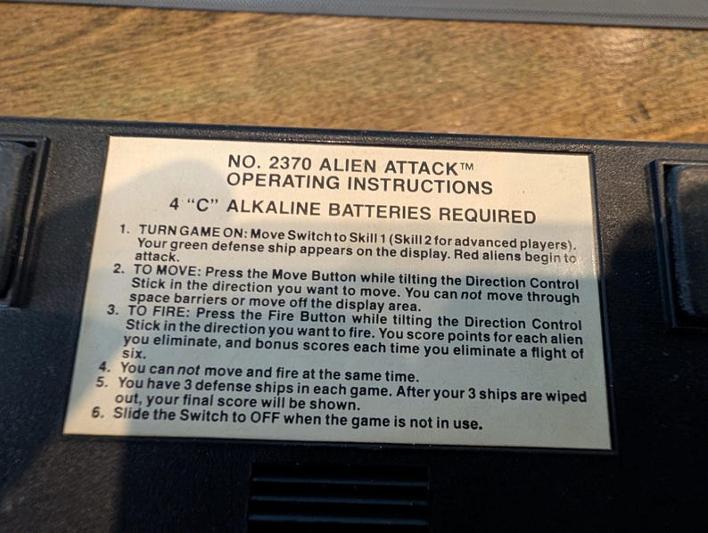 May include: A close-up of a black game console with a white label. The label reads "NO. 2370 ALIEN ATTACK" and includes operating instructions. The text details how to play the game, including moving, firing, and game rules. Requires 4 "C" alkaline batteries.