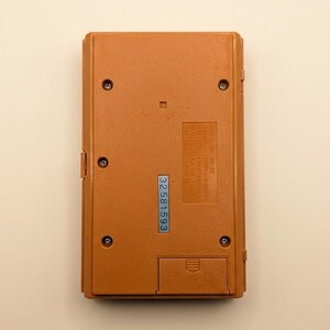 K&ouml;nnte beinhalten: R&uuml;ckseite einer Vintage-Handheld-Spielkonsole in warmem Braun. Sichtbar sind Schrauben, ein Batteriefach und ein blauer Aufkleber mit der Nummer 32581593. Das Ger&auml;t hat eine rechteckige Form.