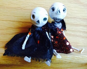 Puede incluir: Dos muñecas esqueleto hechas a mano con caras pintadas. Una muñeca lleva un vestido negro con una superposición de encaje y la otra lleva un vestido de lentejuelas rojo y negro.