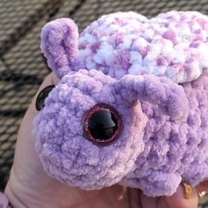 Puede incluir: Un animal de peluche de color morado tejido a crochet con una concha de crochet blanca y morada en la espalda. El animal tiene ojos grandes y brillantes y una nariz rosa.