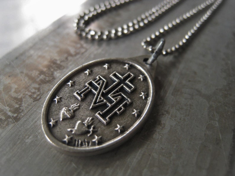 Puede incluir: Un collar de cadena de plata con un colgante de plata que presenta un dise&ntilde;o de cruz estilizado con un coraz&oacute;n y llamas. El colgante tiene forma ovalada y presenta un dise&ntilde;o detallado.