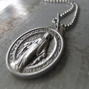 Puede incluir: Un collar de cadena de plata con un colgante de plata que representa a la Virgen Mar&iacute;a. El colgante tiene la inscripci&oacute;n "Oh Maria Sacada Concebida, Ruega Por Nosotros" alrededor del borde.