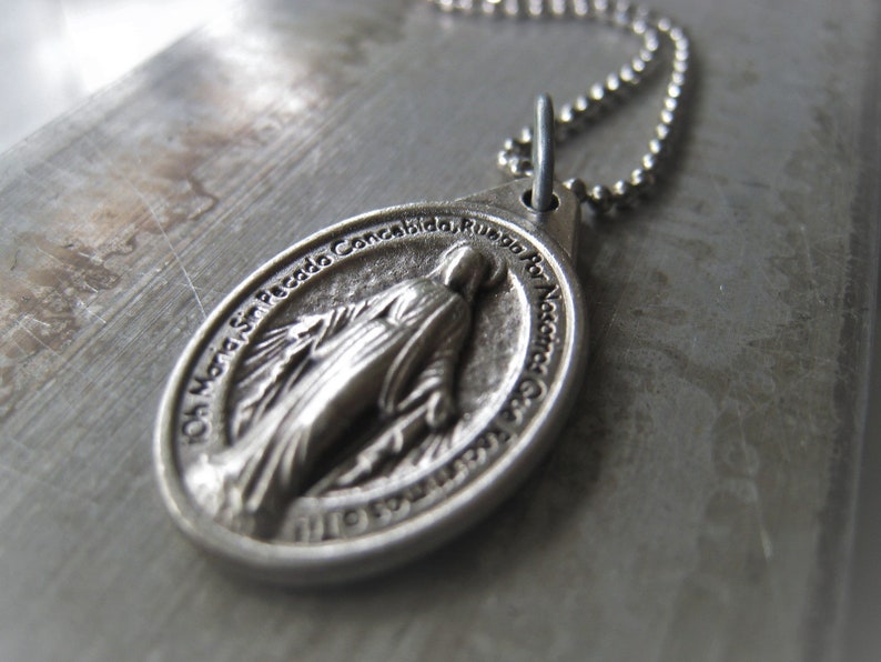 Puede incluir: Un collar de cadena de plata con un colgante de plata que representa a la Virgen Mar&iacute;a. El colgante tiene la inscripci&oacute;n "Oh Maria Sacada Concebida, Ruega Por Nosotros" alrededor del borde.