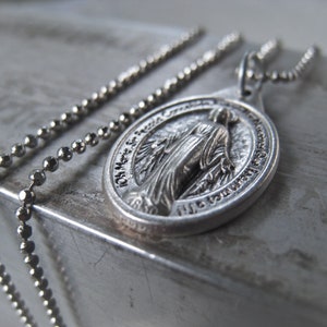 Puede incluir: Un collar de cadena de plata con un colgante de plata que presenta una imagen religiosa de la Virgen Mar&iacute;a. El colgante tiene las palabras "Oh Mar&iacute;a concebida sin pecado ruega por nosotros que recurrimos a ti" inscritas alrededor de la imagen.