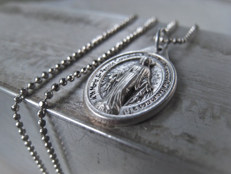 Puede incluir: Un collar de cadena de plata con un colgante de plata que presenta una imagen religiosa de la Virgen Mar&iacute;a. El colgante tiene las palabras "Oh Mar&iacute;a concebida sin pecado ruega por nosotros que recurrimos a ti" inscritas alrededor de la imagen.