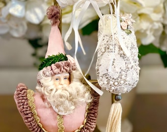 May include: Three Christmas ornaments hanging on a decorative gold stand. The ornaments include a pink Santa Claus, a white lace doll, and a white lace ornament with pearls and a tassel. The background features white flowers.