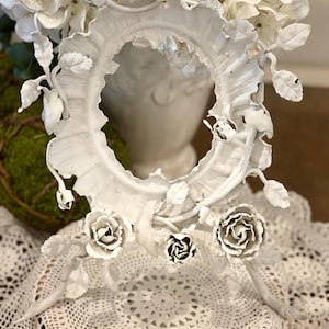 May include: White, ornate, oval-shaped picture frame with rose and leaf details. The frame is decorated with several white roses and leaves. The frame is placed in front of a white vase filled with white flowers.