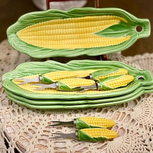 May include: A set of ceramic corn-shaped serving dishes and corn-on-the-cob forks. The dishes are green with yellow corn detailing. The forks have silver tines and corn-shaped handles. The set is designed for serving corn on the cob.
