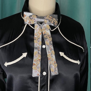May include: A black satin shirt with white piping and arrow-shaped embroidery. A silver bow tie with intricate gold and pink embroidery and embellishments is centered on the collar. The shirt has white buttons.