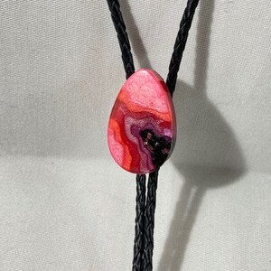 May include: A teardrop-shaped bolo tie pendant with a marbled design in shades of pink, red, and black. The pendant is suspended from a black braided cord. This accessory adds a touch of artistic flair.