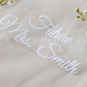 Pode incluir: Um véu branco transparente com o texto bordado "Mrs. Future Smith" em escrita elegante. Delicados detalhes florais são colocados perto da borda superior. O véu é provavelmente para um casamento ou evento nupcial.