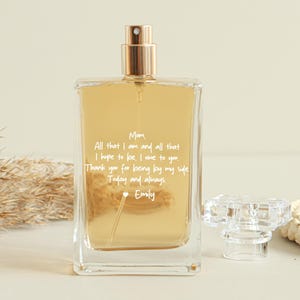 Puede incluir: Un frasco de perfume rectangular con tapa dorada y cristal transparente. El frasco contiene un líquido dorado y tiene texto blanco que dice "Mom, All that I am and all that I hope to be, I owe to you. Thank you for being by my side Today and always Emily."