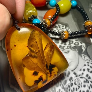 May include: A pendant necklace featuring a large, amber-coloured resin pendant. The pendant contains a preserved leaf and yellow floral elements. The necklace also has a beaded strand with orange, yellow, and turquoise beads.
