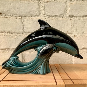 Peut inclure: Une sculpture de dauphin en céramique noire et turquoise brillante. Le dauphin est arqué, sautant au-dessus d'une vague turquoise. La sculpture est posée contre un mur de briques claires et une surface en bois.