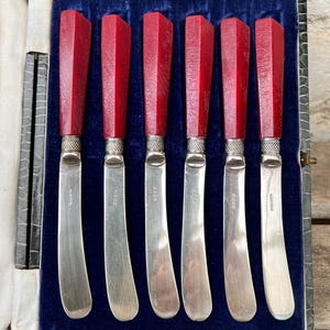 May include: A set of six silver-plated butter knives with faceted, deep red handles and textured silver ferrules. Each knife features a curved blade with engraved hallmarks, presented in a rectangular case lined with dark blue velvet fabric.