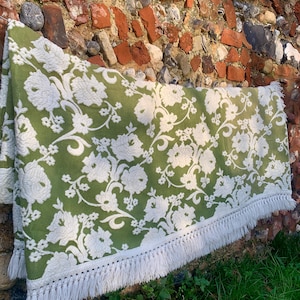 May include: A green and white floral patterned blanket with a fringe trim. The blanket features a repeating pattern of white flowers and vines on a green background. The fringe is white.