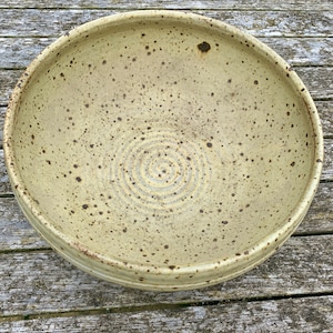 May include: A large, round, ceramic bowl with a speckled, light yellow glaze and brown speckles. The bowl has a slightly raised rim and a concentric circle pattern in the centre.