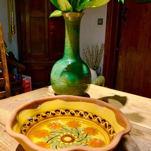 Może przedstawiać: Ozdobna ceramiczna miska z żółtym wnętrzem i kwiatowym wzorem, na drewnianym stole. Wysoki zielony wazon z zielonymi liśćmi stoi za miską, tworząc naturalną atmosferę.