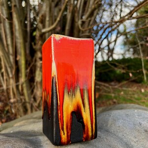 May include: A rectangular ceramic vase with a vibrant color scheme. The top is a glossy red, transitioning to yellow and black at the base. The vase is displayed outdoors on a gray rock, with a blurred natural background.