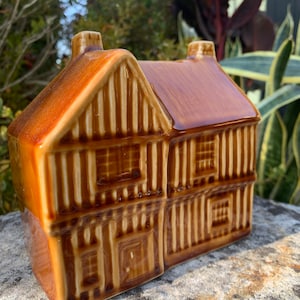 Puede incluir: Una figurita de casa de cerámica marrón con molduras blancas y una fachada detallada. La casa tiene un techo a dos aguas y dos chimeneas.