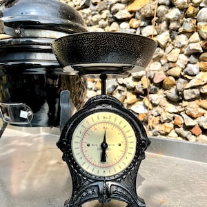 May include: A black mechanical kitchen scale with a round, textured black weighing bowl. The circular dial features a cream-colored face with black and red markings, displaying measurements in both kilograms and pounds. The text reads Made Sweden.